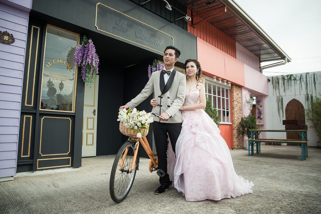 bride and groom on a bicycle in full wedding attire. Groom in grey suit and bride in a plush pink dress