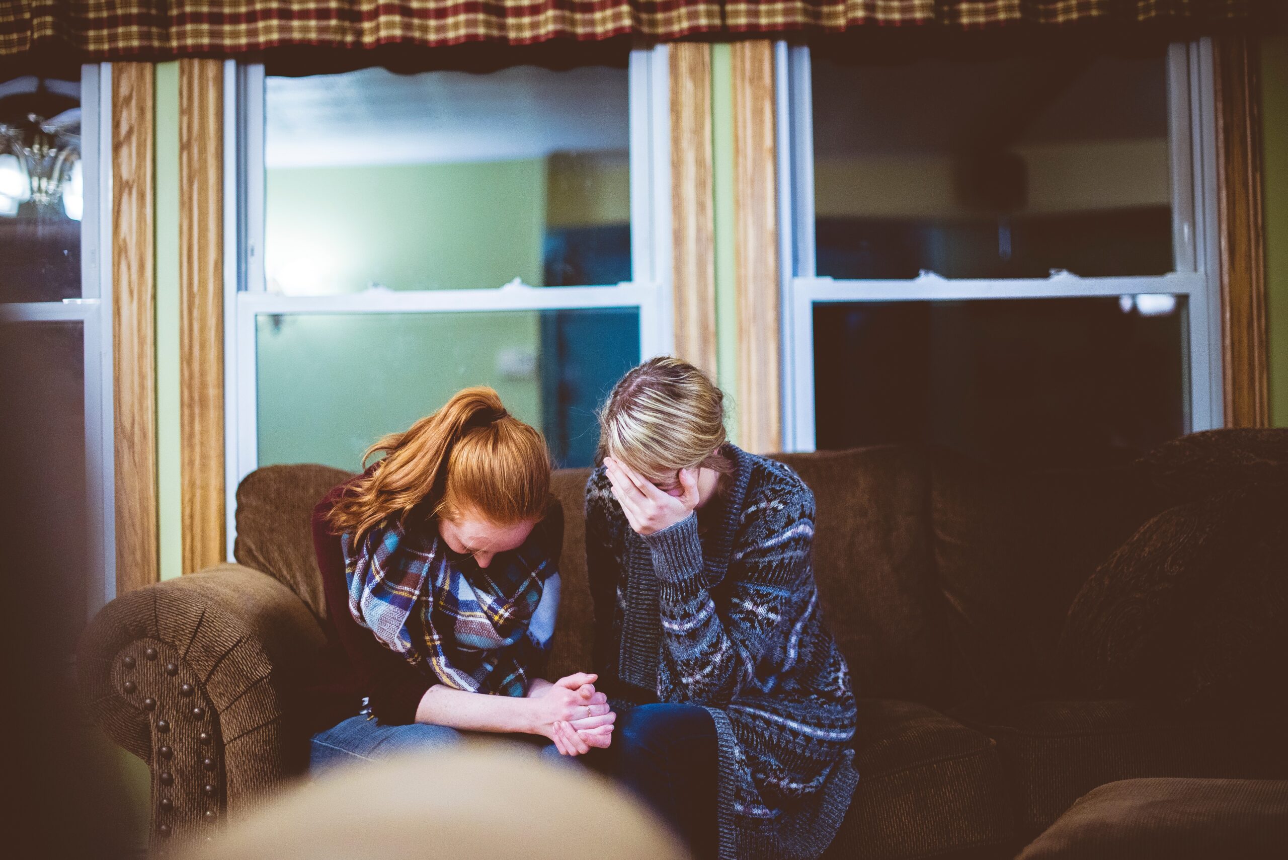 two women sat on sofa with heads in heands and holding each others hand
