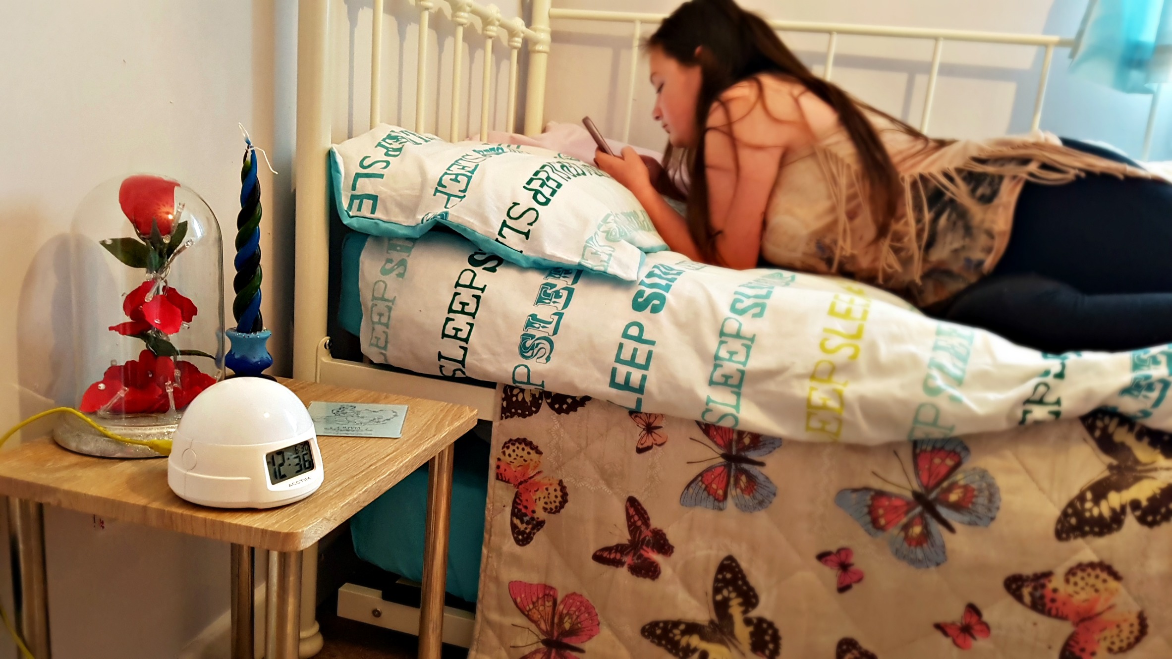 girl lying on a bed on a duvet saying sleep next to a bedside table with a rose ornament and alarm clock