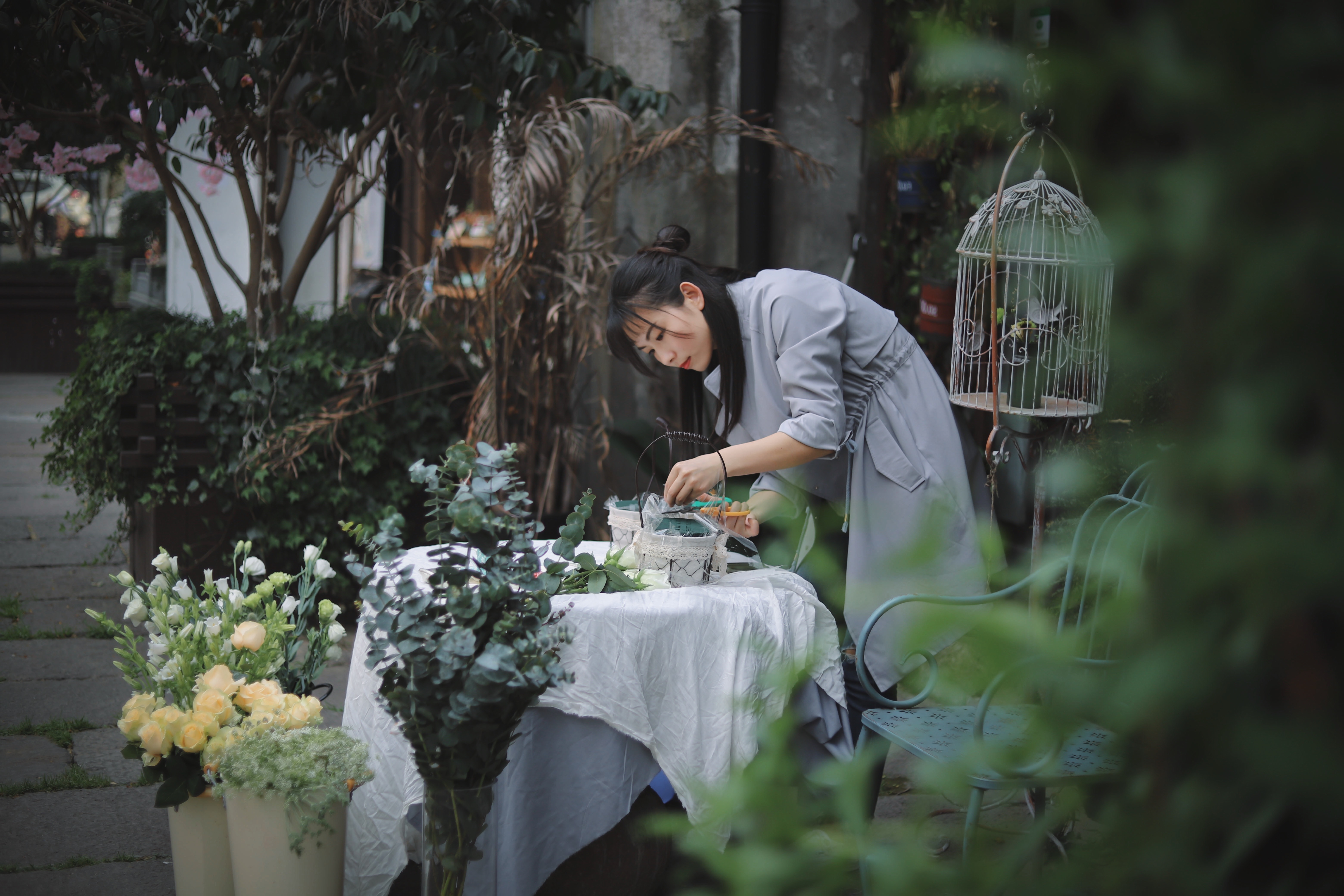 woman tending to plants on a table with flowers around her and a birdcage in the background