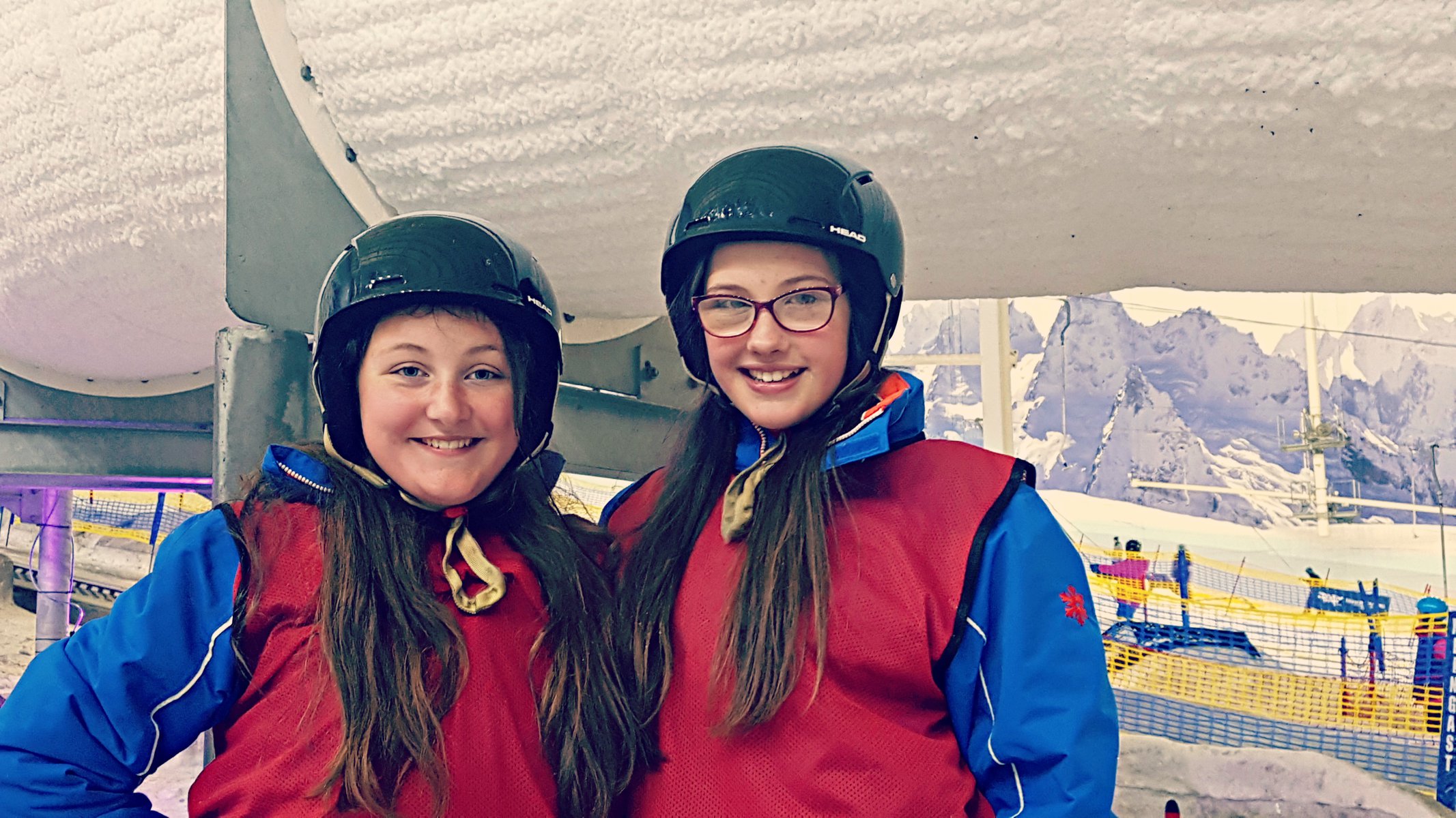 2 girls in helmets and ski wear a an indoor ski park