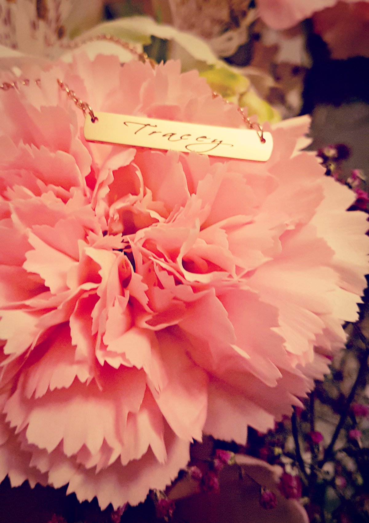 the personalised rose gold bar necklace from inscripture with the name tracey lying on a pink flower