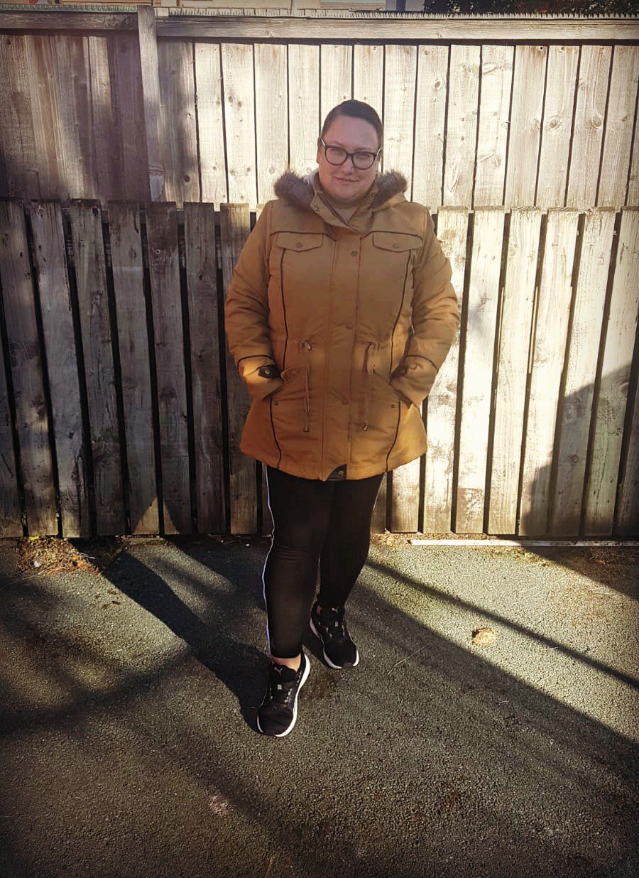 woman standing in a shaded garden with a mustard coat on with a fur lined hood