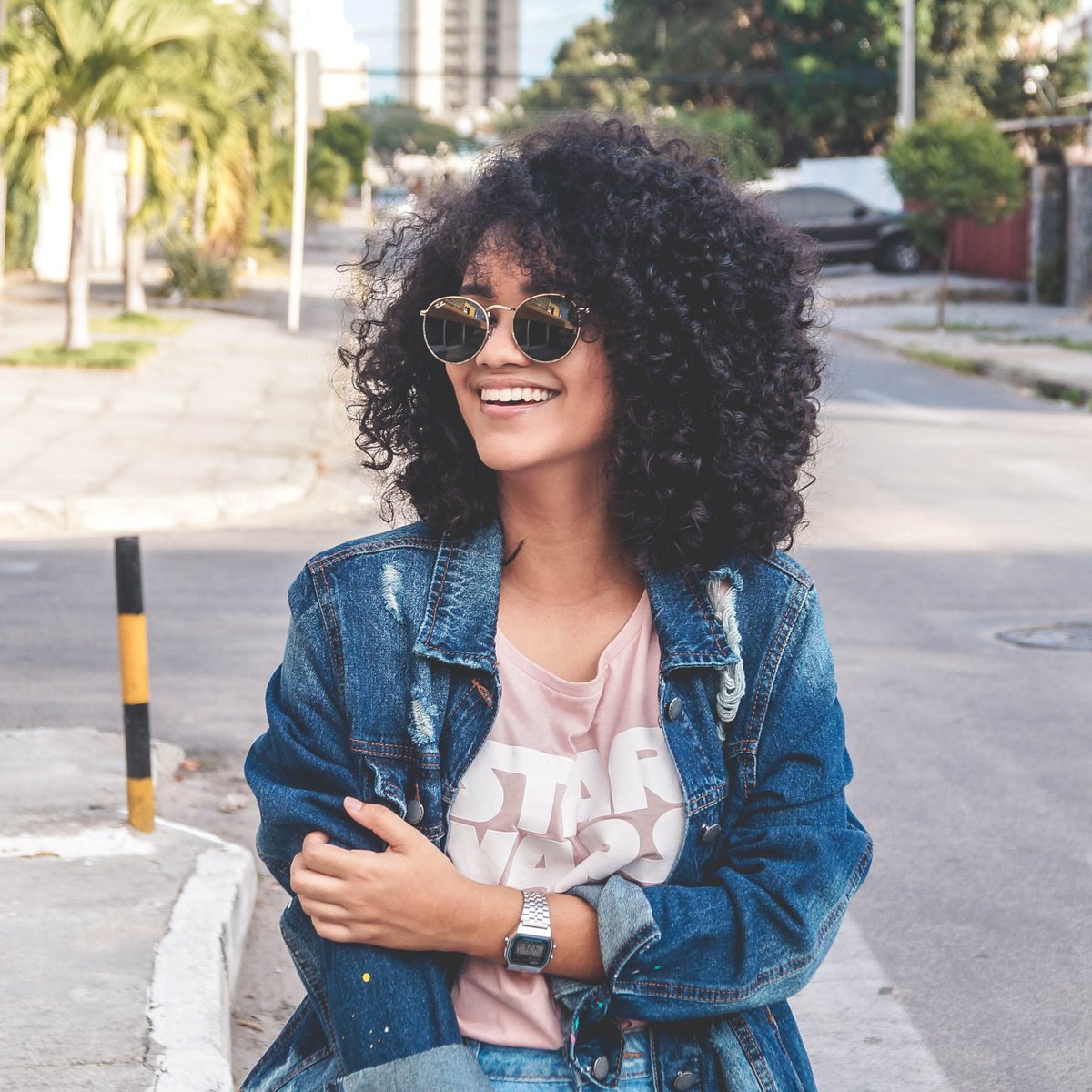 girl with curly hair wearing sunglasses standing in road with a denim jacket on, arms folded in front of her with a star wars tshirt showing under the jacket