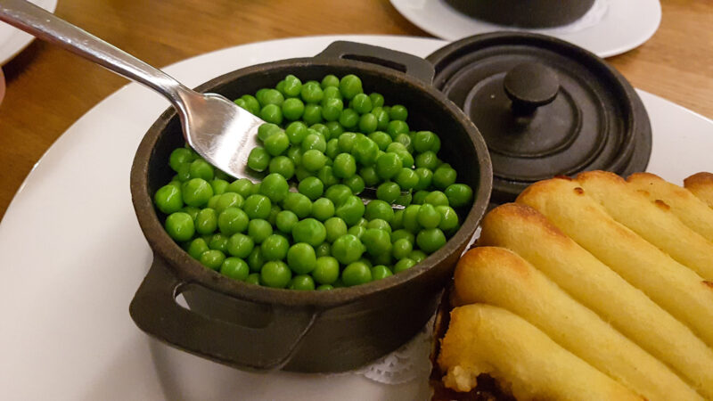 a black dish with garden peas next to a shepherds pie on a white plate