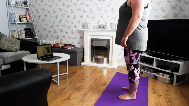 a woman in sportswear standing on a purple fitness mat with a laptop in view on living furniture