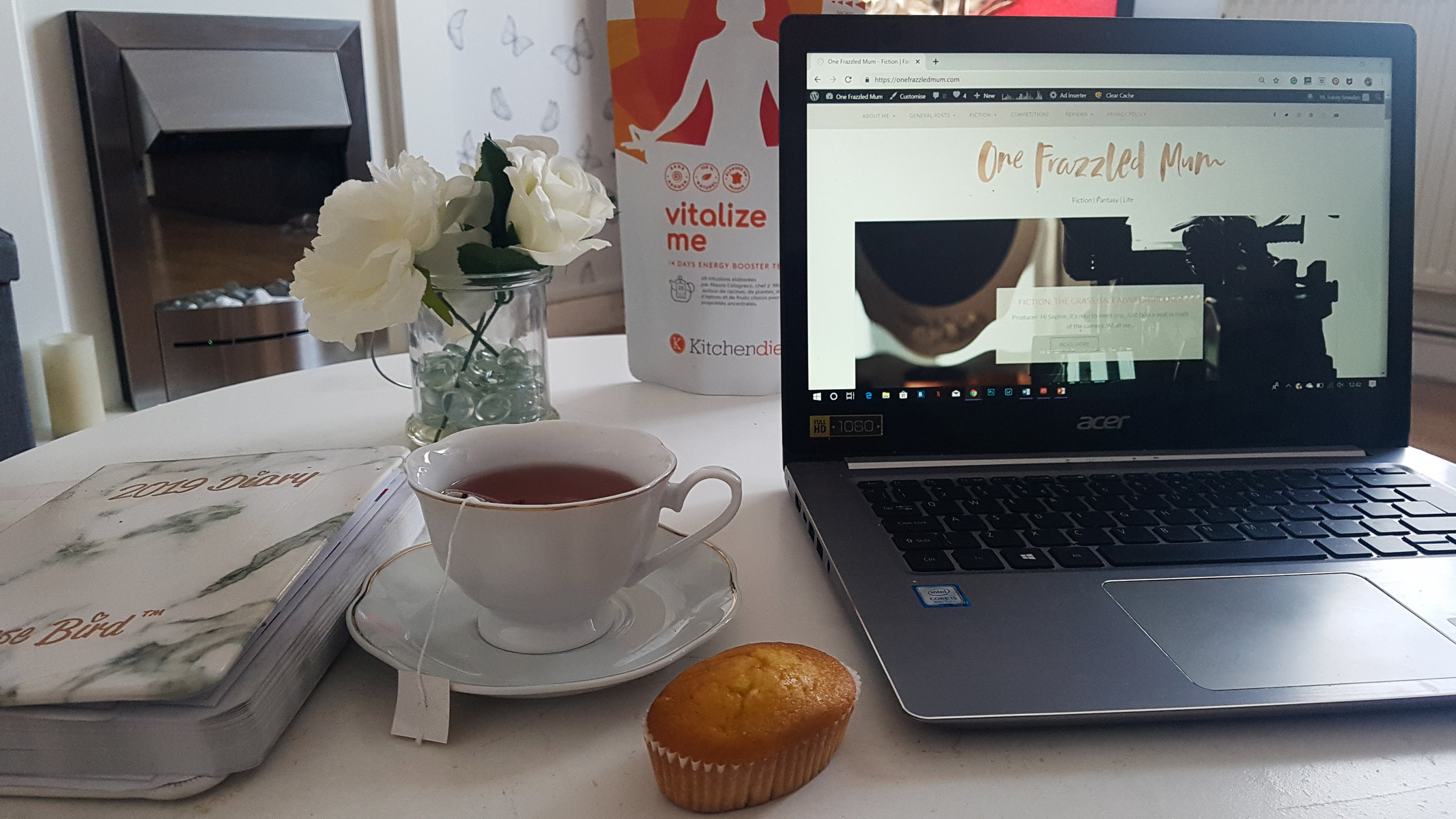 an open laptop on a white coffee table with a diary, small muffin and tea in a white china cup and saucer