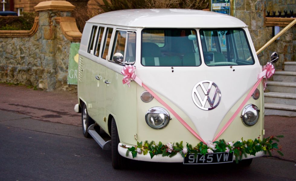 a white and beige vw camper van with pink ribbon on the front and flowers across the bumper