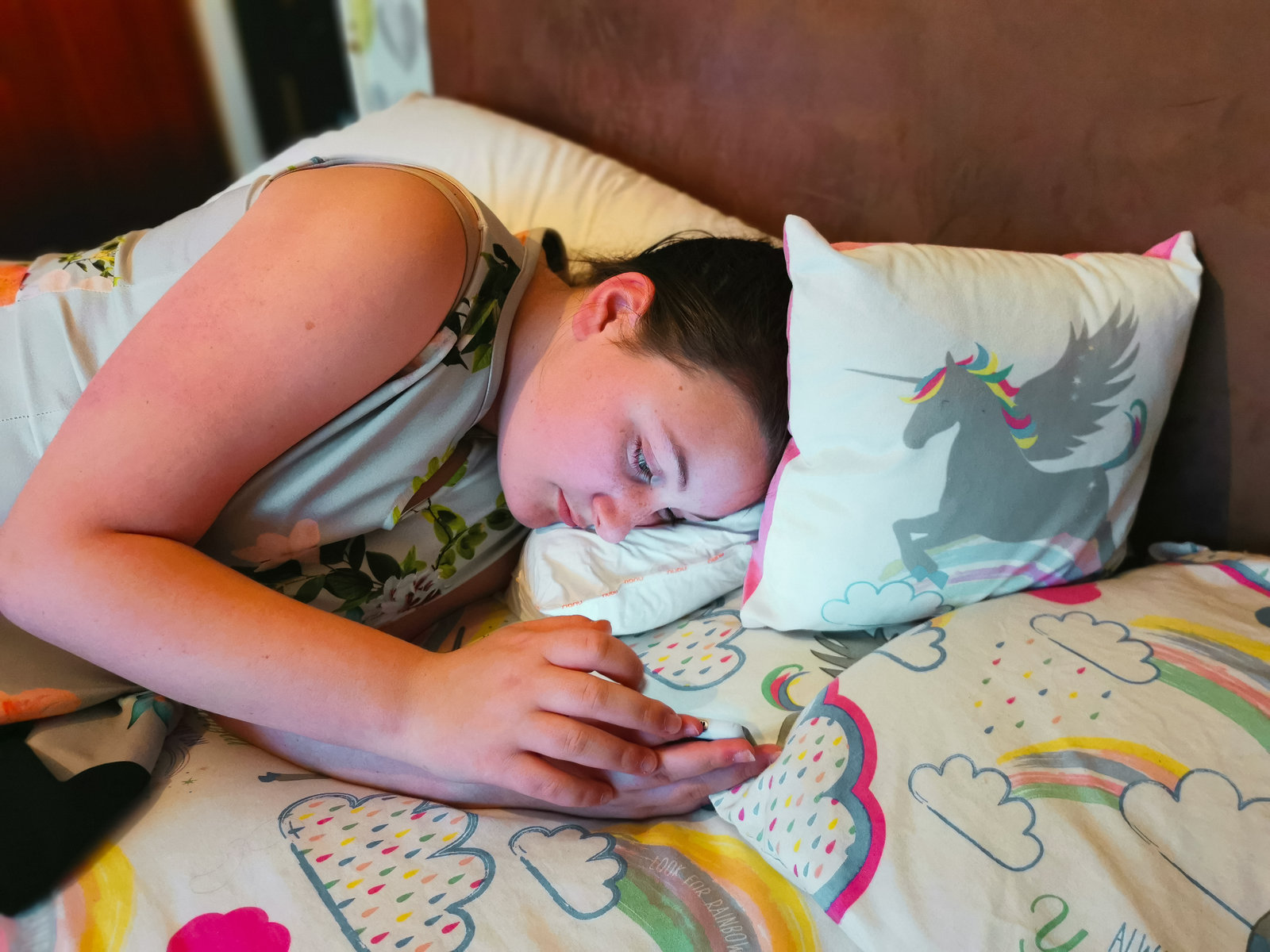 young girl lying on a bed using a phone with a unicorn pillow by her head