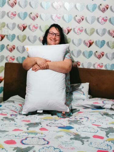 a woman wearing glasses kneeling on a bed holding a nanu pillow