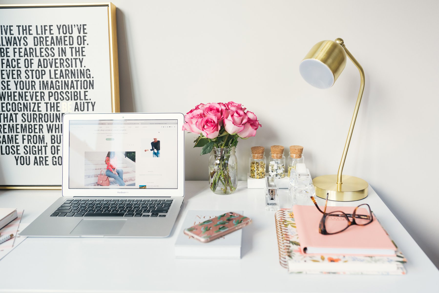 a white desk with staionery, magazines and framed pictures around an open laptop
