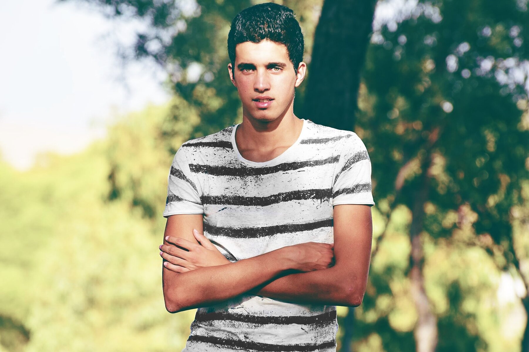 young boy with black hair wearing a stripey top standing arms crossed in a grassy area
