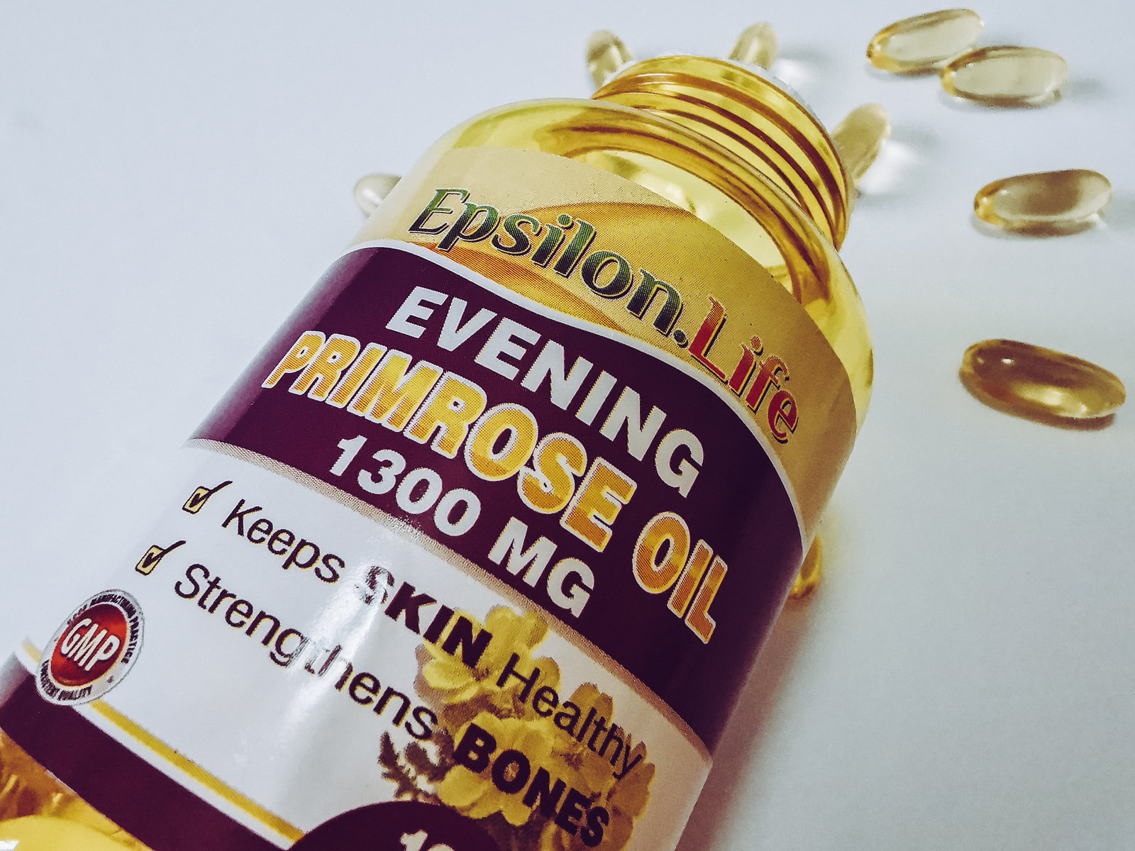 an open bottle of epsilon life evening primrose oil capsules with the capsules scattered on a white background