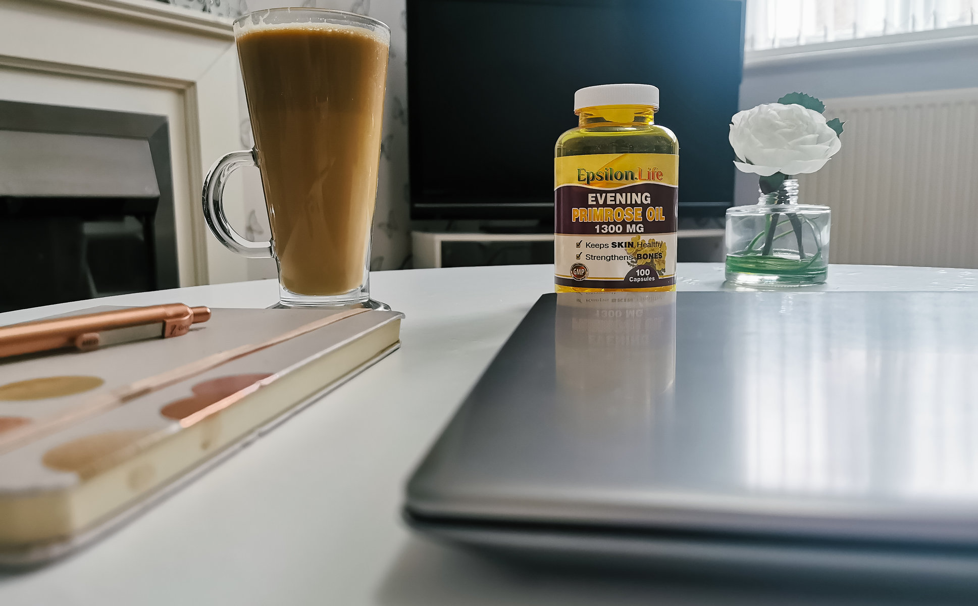 a table set up with a closed laptop, diary, pen, flower, coffee and a bottle of evening primrose oil capsules