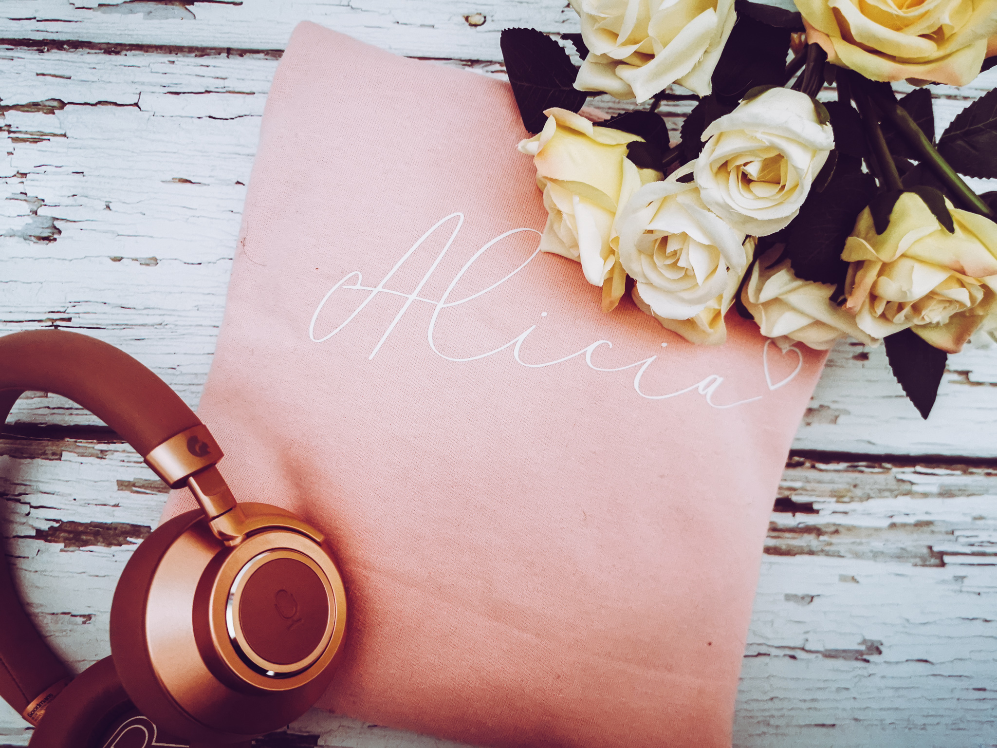 flatlay of a personalised pink jumper with rose gold headphones and artificial flowers in the shot