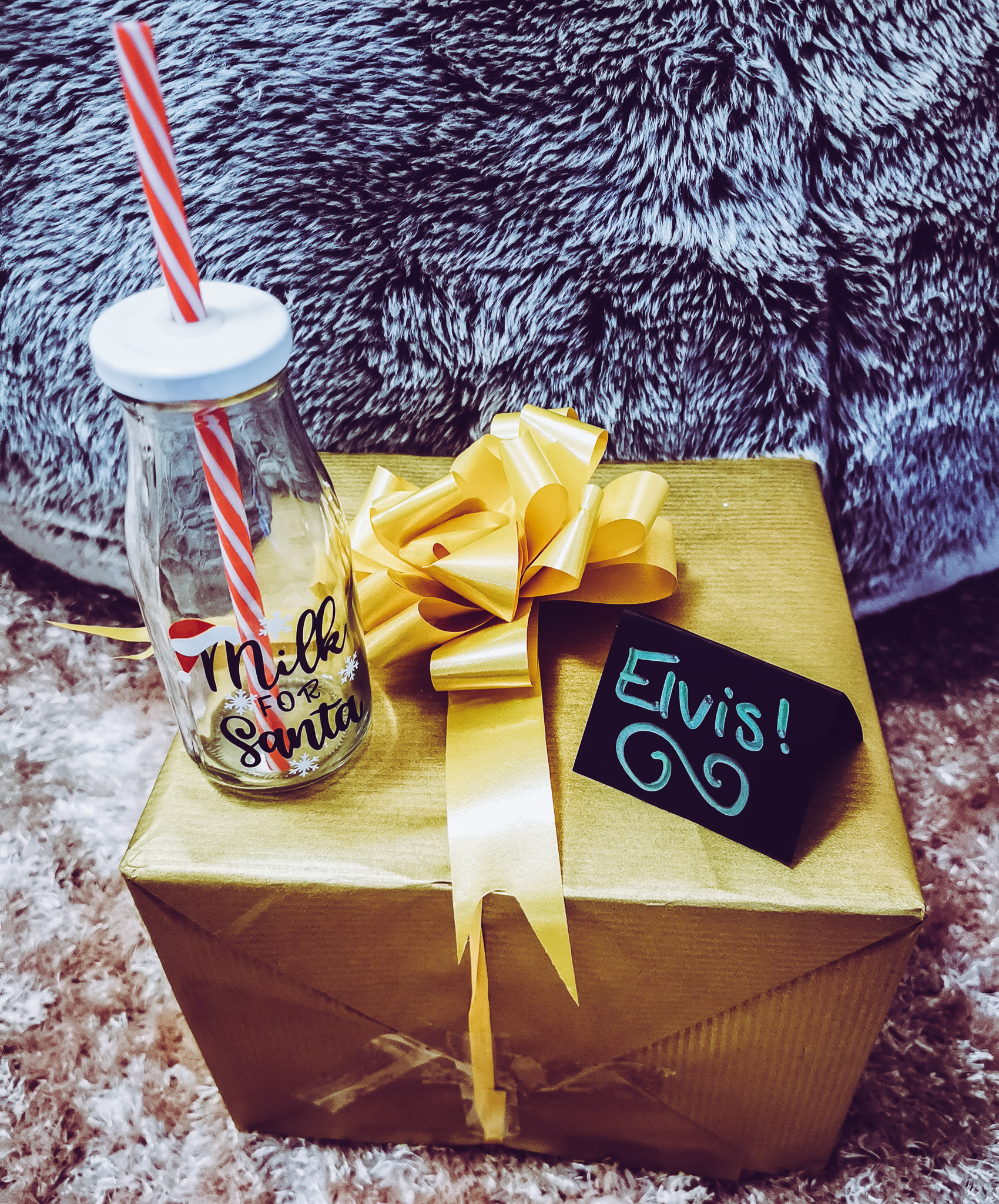 a gold gift wrapped box with a bow and a bottle for milk for santa and a name card