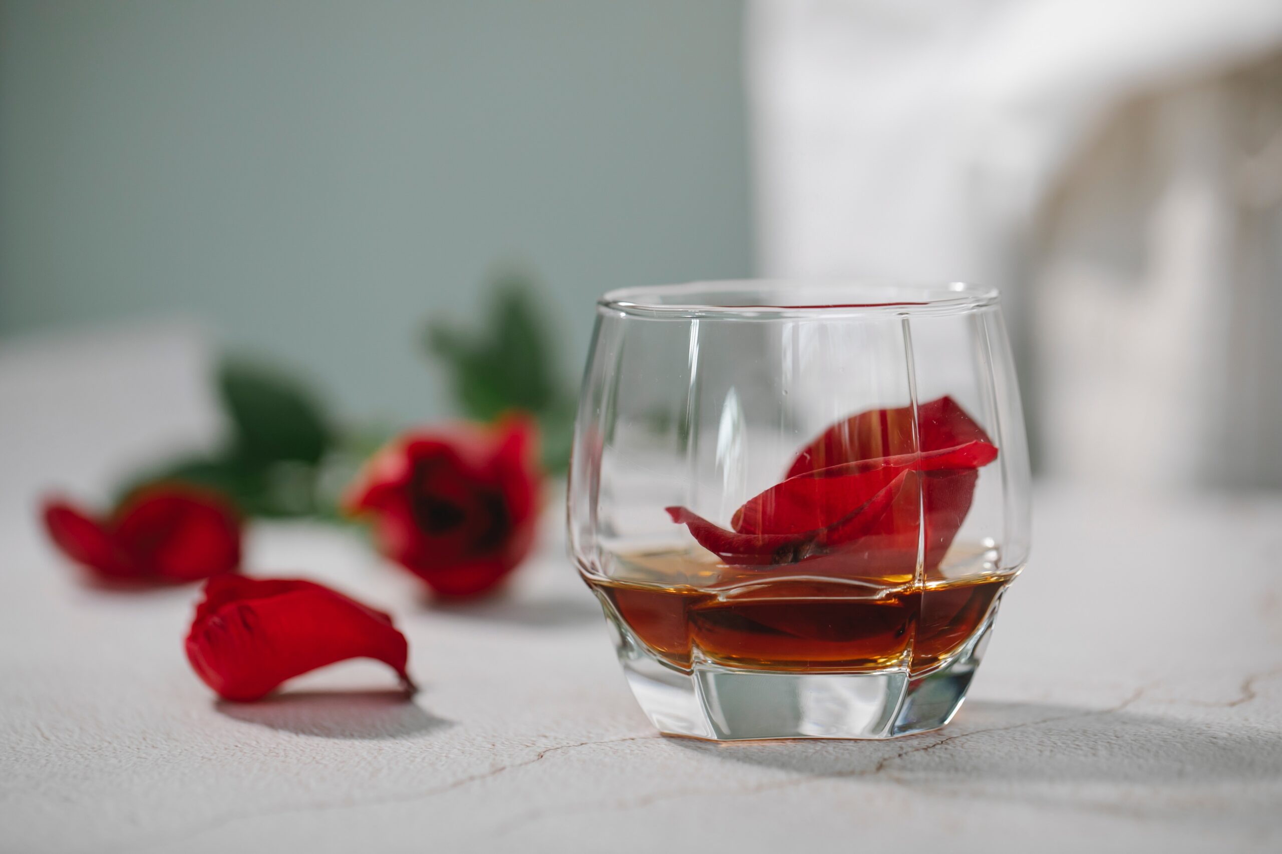 rose petals on a countertop next to a glass with alcohol in it and one rose petal in the glass