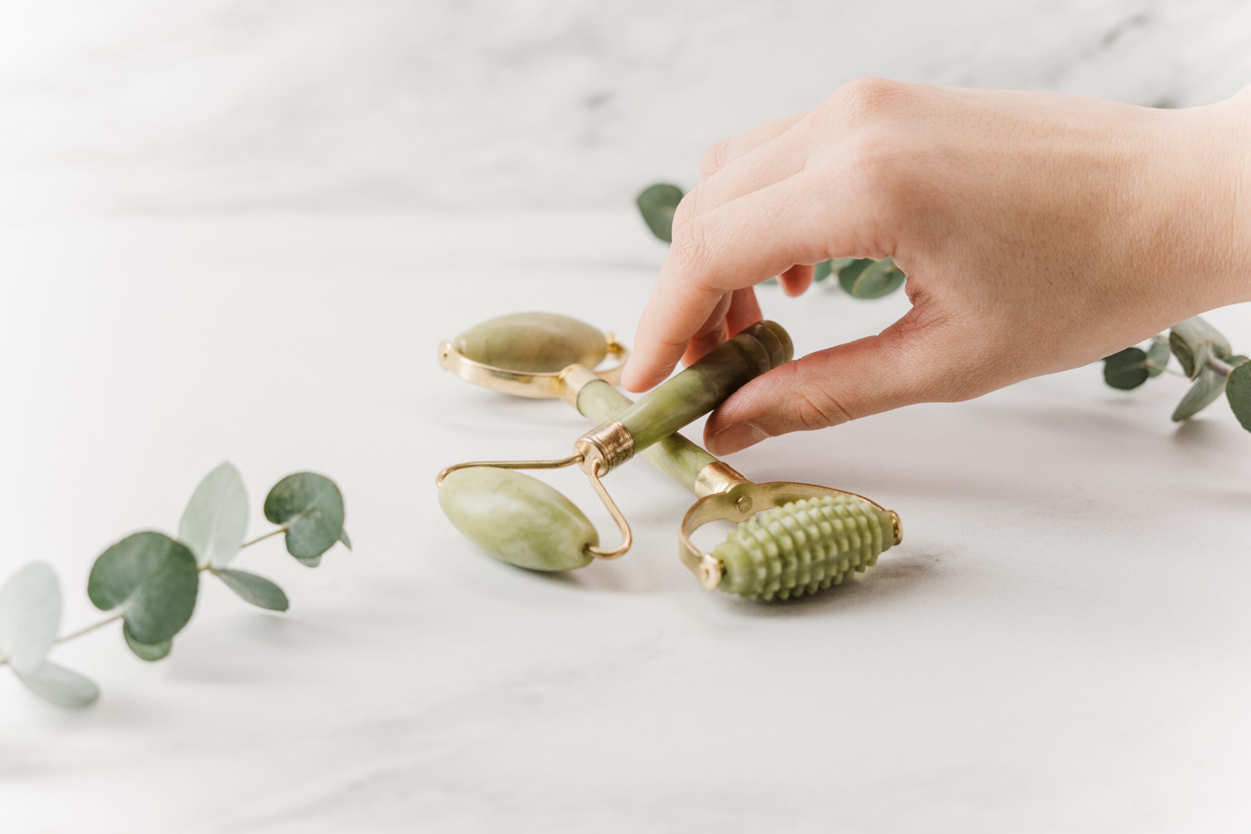 a hand picking up a jade facial roller from a white marble countertop