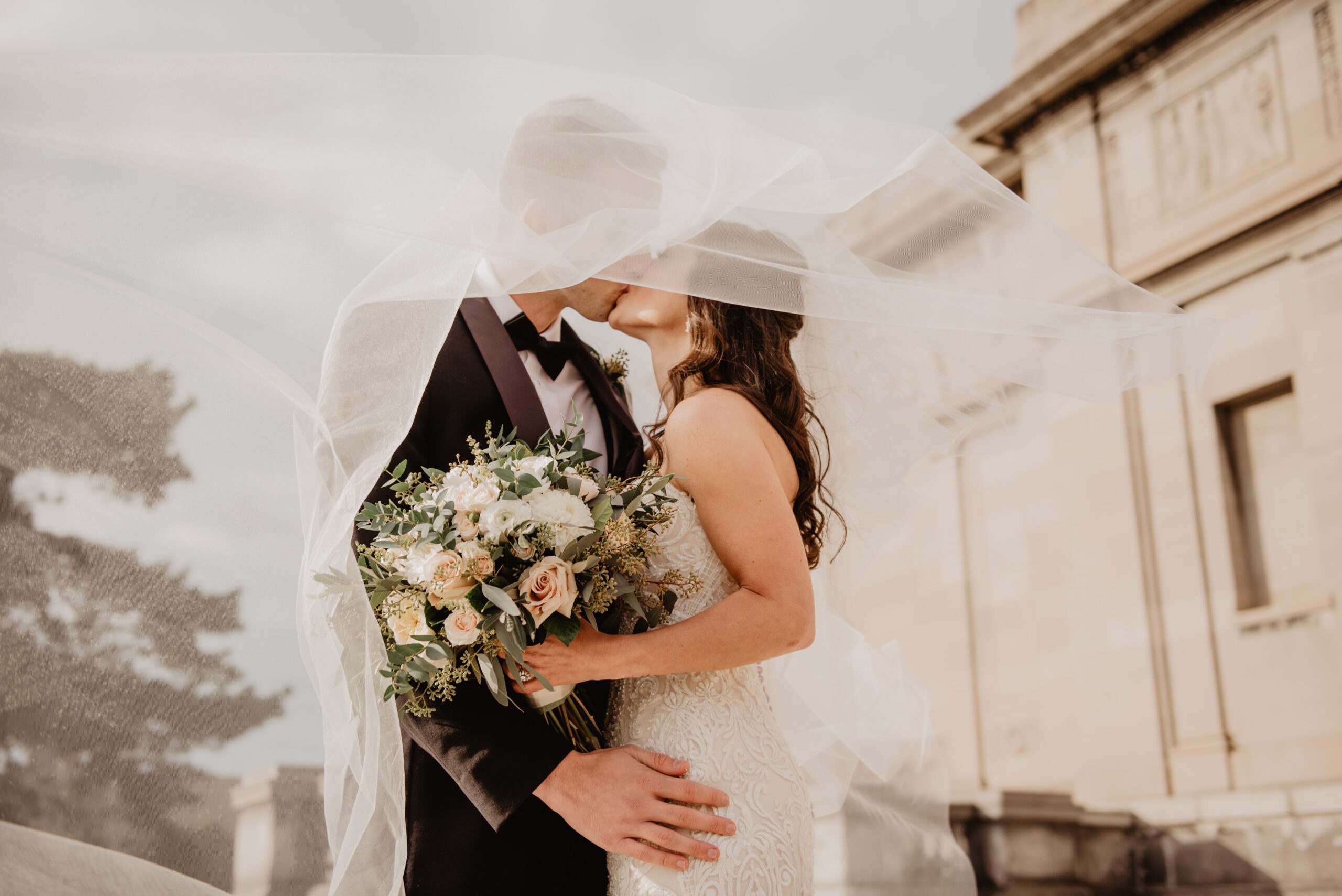 bride and groom under a veil kissing holding a wedding bouquet
