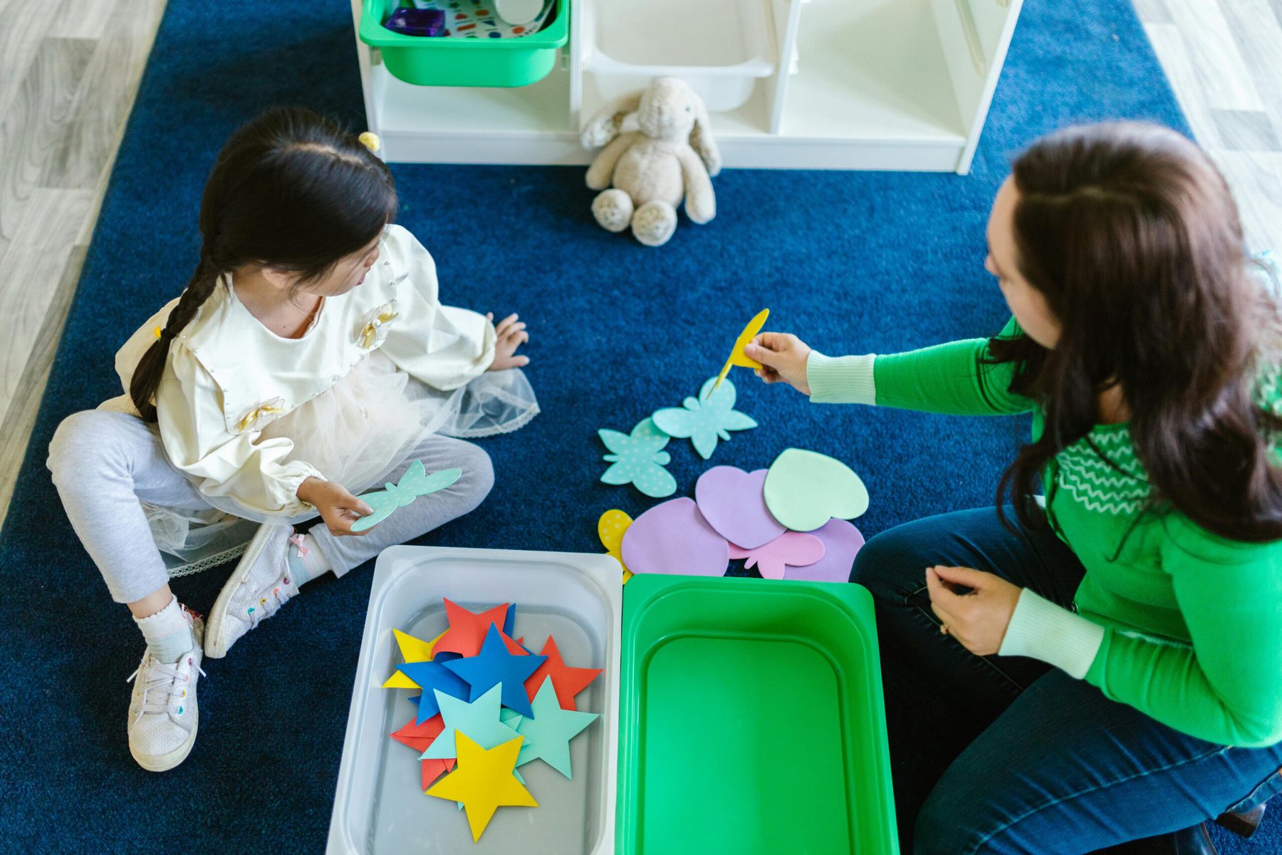 a woman and a child sitting on a blue rug with an open box of craft supplies inbetween them