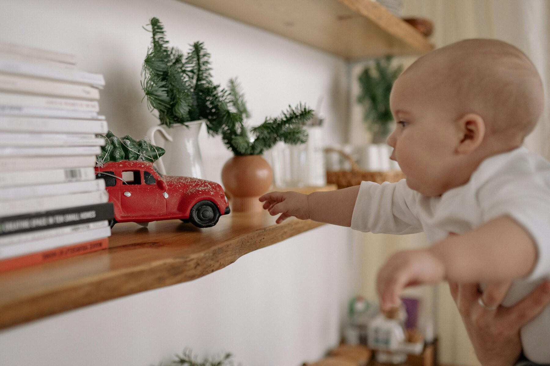 a baby wearing white bing held up as they reach for a red toy on shelf