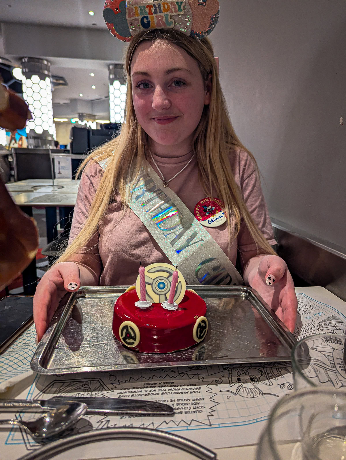 a young girl wearing happy birthday styled minnie mouse ears and a birthday sash is wearign a pink tshrt and holdig a birthday cake with marvel stylings and a red glaze in Pyms Kitchen Disneyland Paris