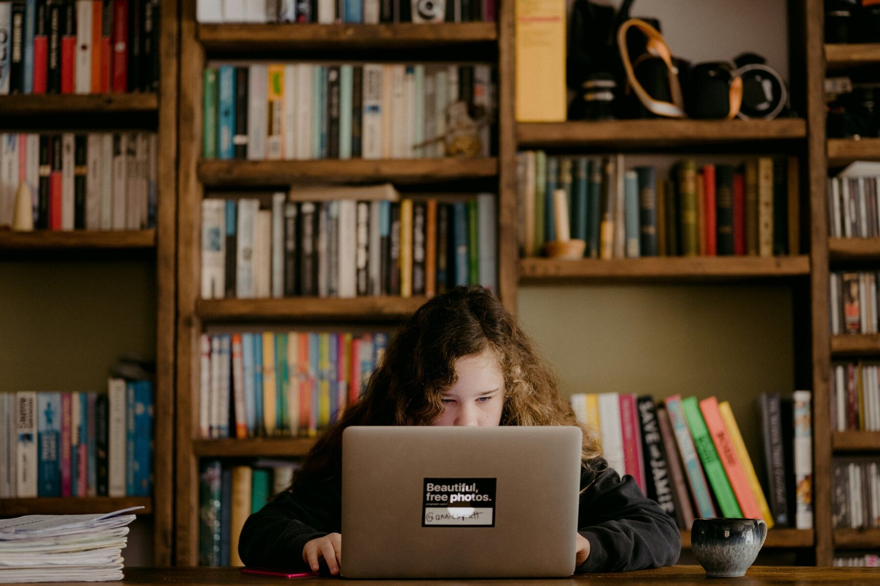 teen with long dark curly hair hunched behid a silver laptop with shelves of books in the background