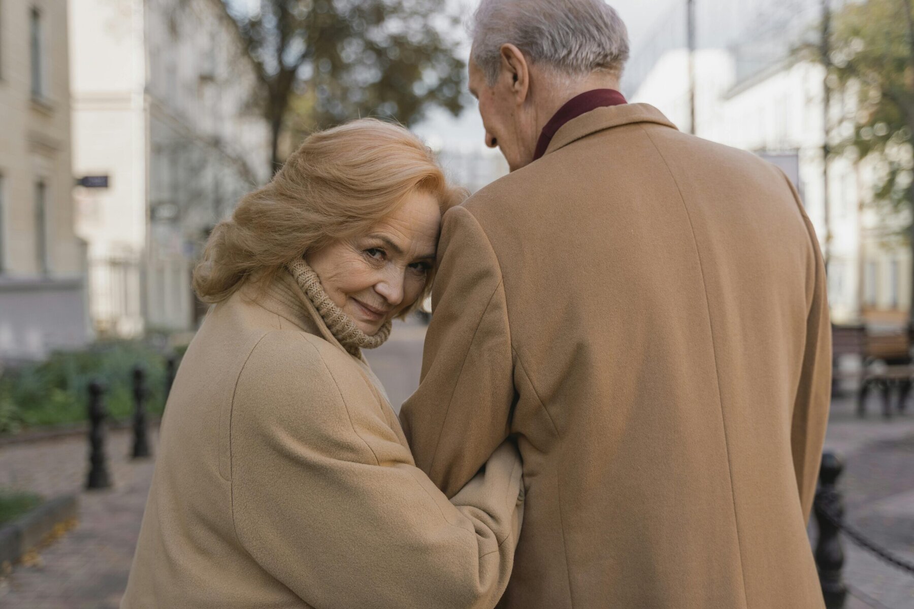 An older couple walking arm-in-arm down a quiet street, dressed warmly, looking content and independent — illustrating the idea of ageing in place with dignity and companionship.