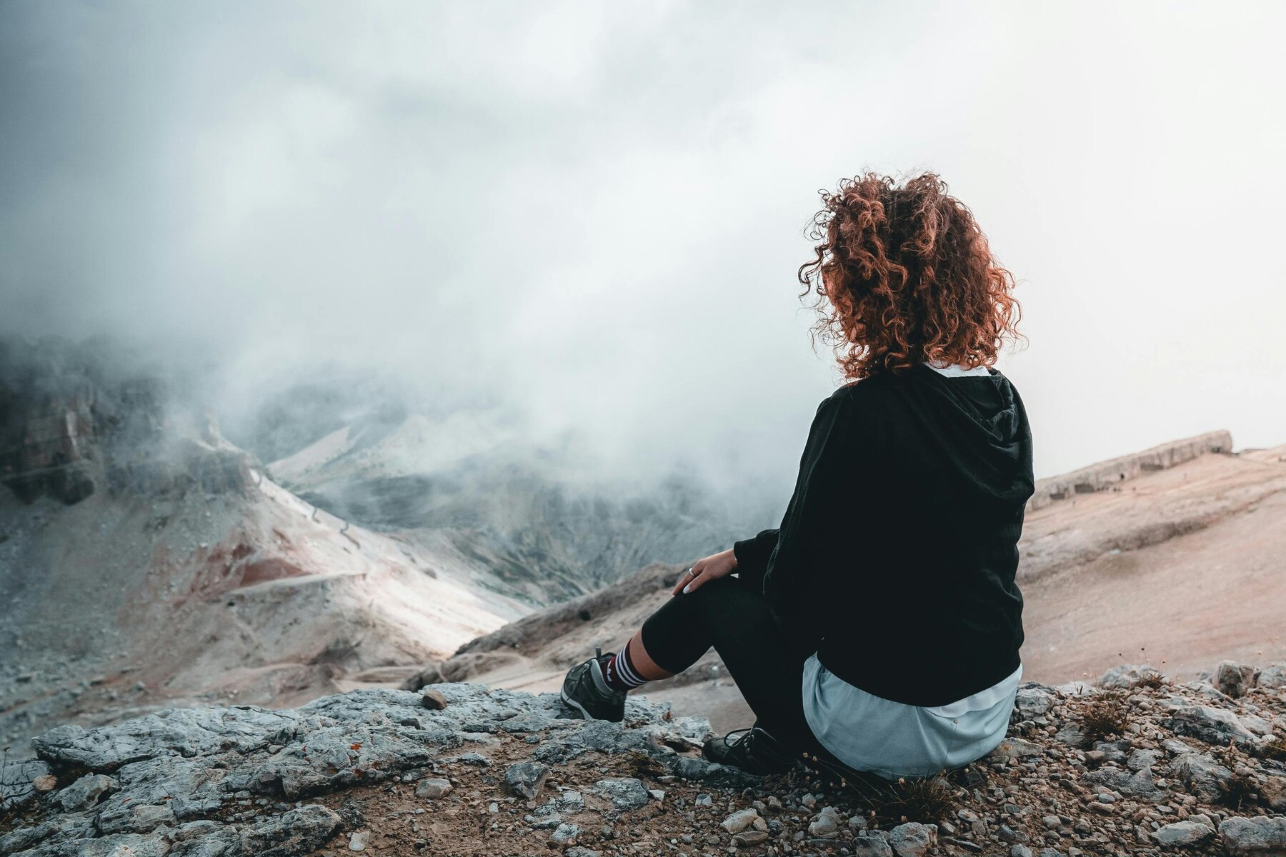 Woman sitting peacefully on a mountainside, looking out over the landscape, taking a quiet moment to breathe and reset.