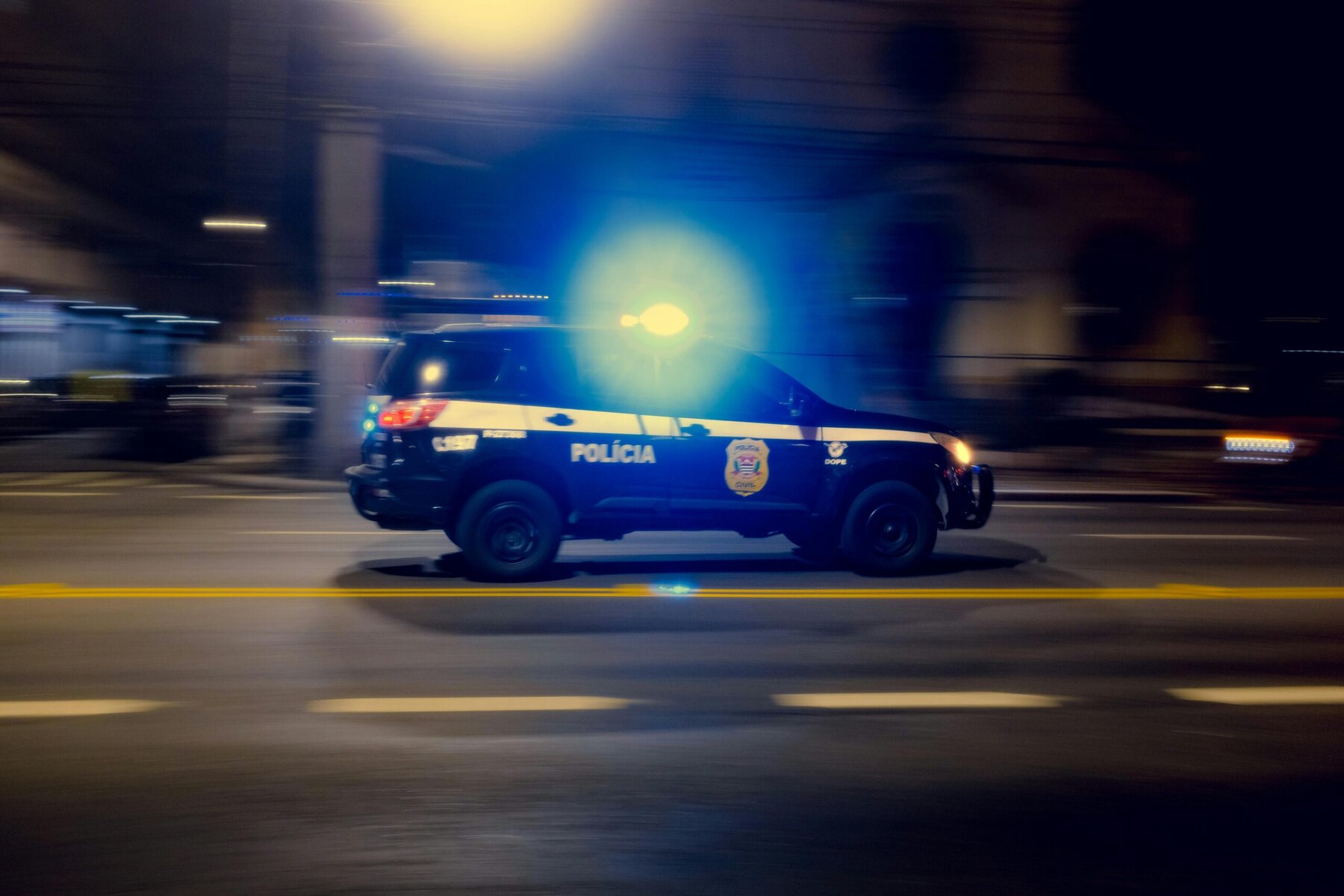 “A police car speeding along a dark urban road at night, its flashing blue and red lights casting motion blur against city streetlights.”