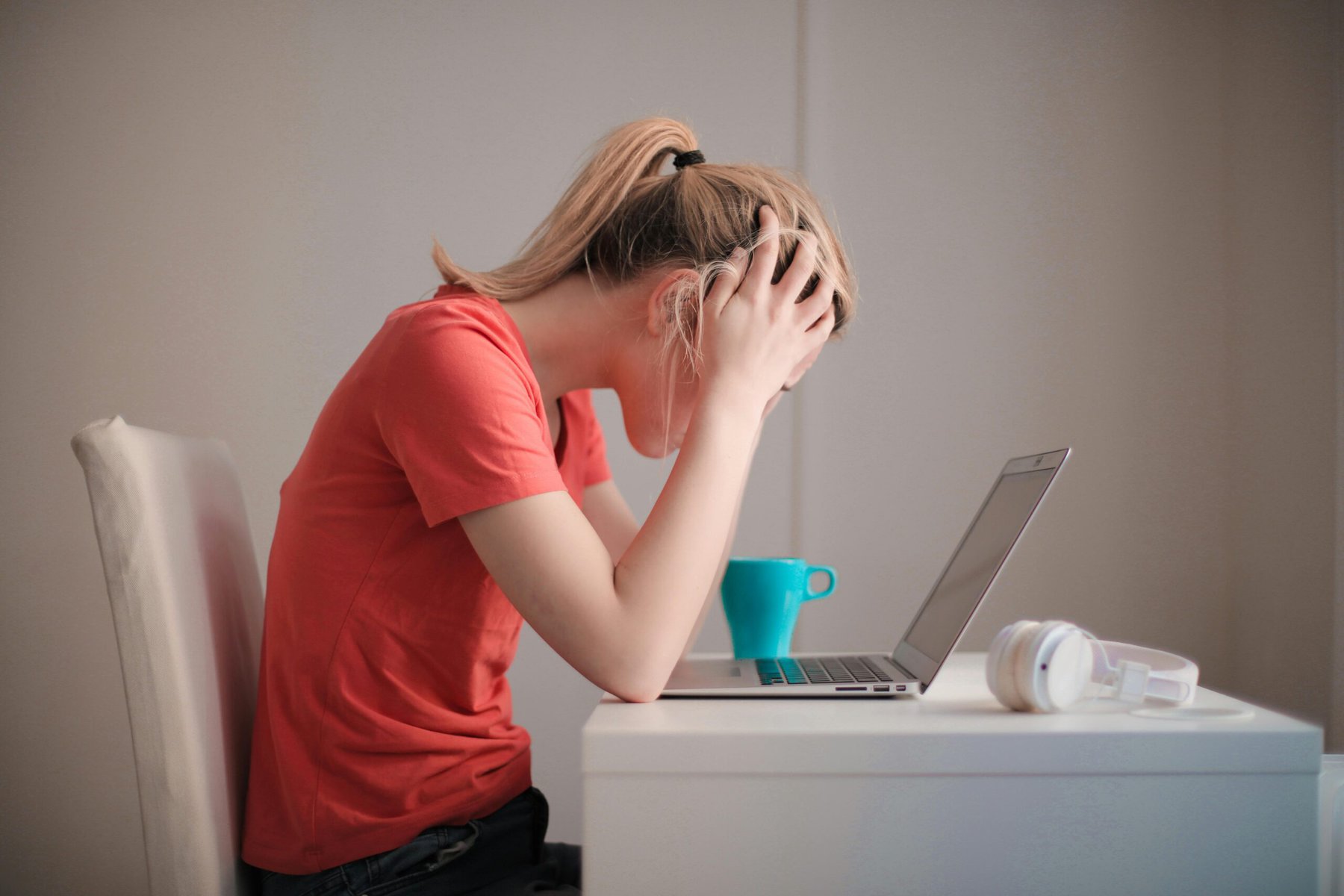 Young woman in a red T-shirt sitting indoors at a table, looking worried or stressed while using a laptop — a coffee mug and headphones sit nearby.”