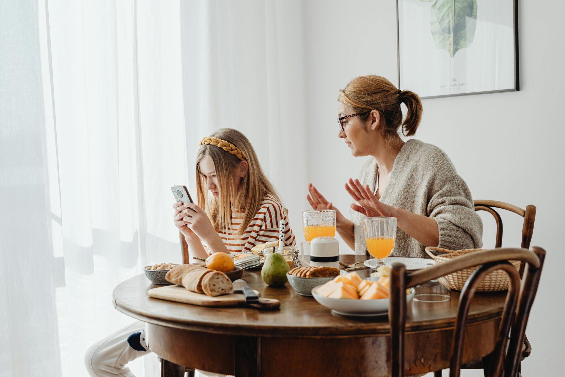 A mother looking disapprovingly at her teenage daughter's phone during breakfast, illustrating the challenge of teaching good manners and screen etiquette at home